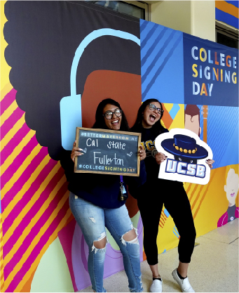Two students posing in front of backdrop holding signs with college names