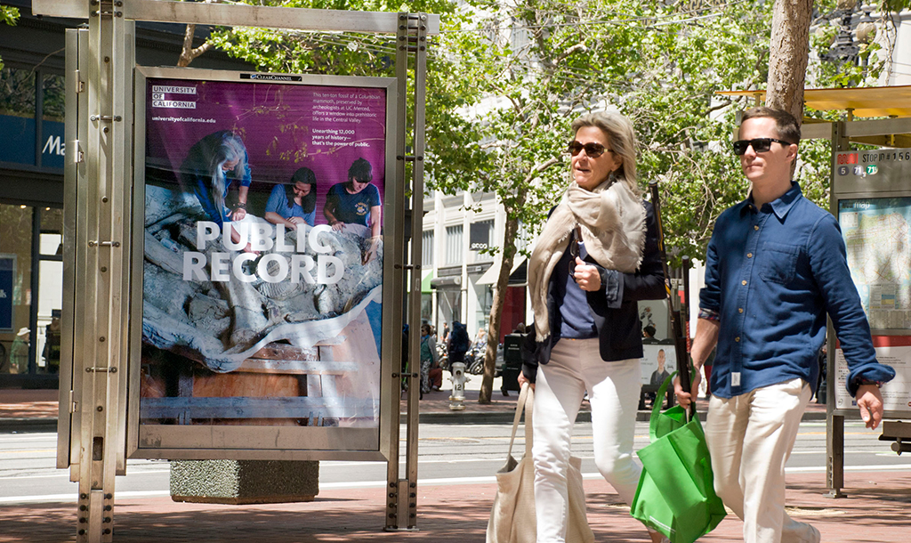 Bus shelter with poster of a professor and students looking at fossils with the words Public Record